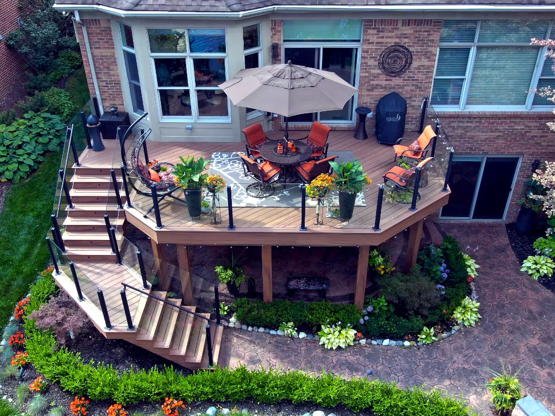 Drone aerial of a curved Trex composite deck with glass railing, built-in seating area and landscaped stone patio below