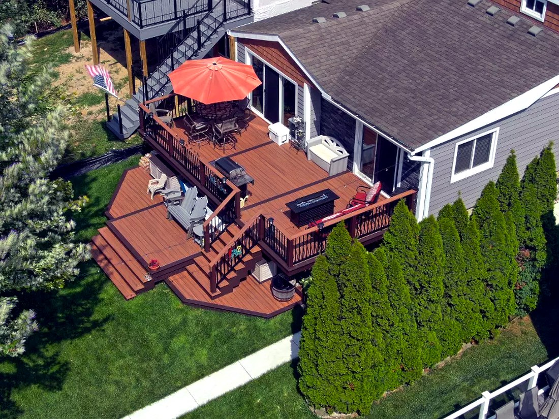 Drone aerial of a multi-level Trex composite deck with fire pit table and umbrella, overlooking a landscaped backyard